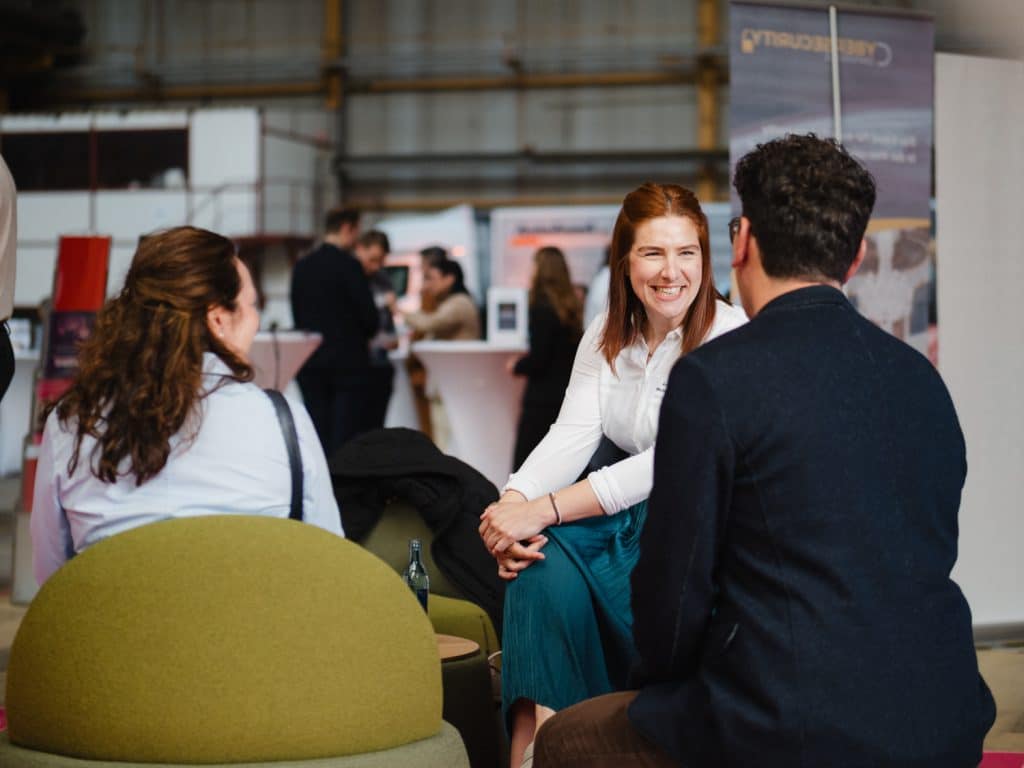 sympathische Frau auf einem Event während einenm angeregten Gespräch. Die Umgebung ist eine große moderne Messehalle, die viel Wärme vermittelt. Im Hintergrund sind weitere Stände zu sehen.