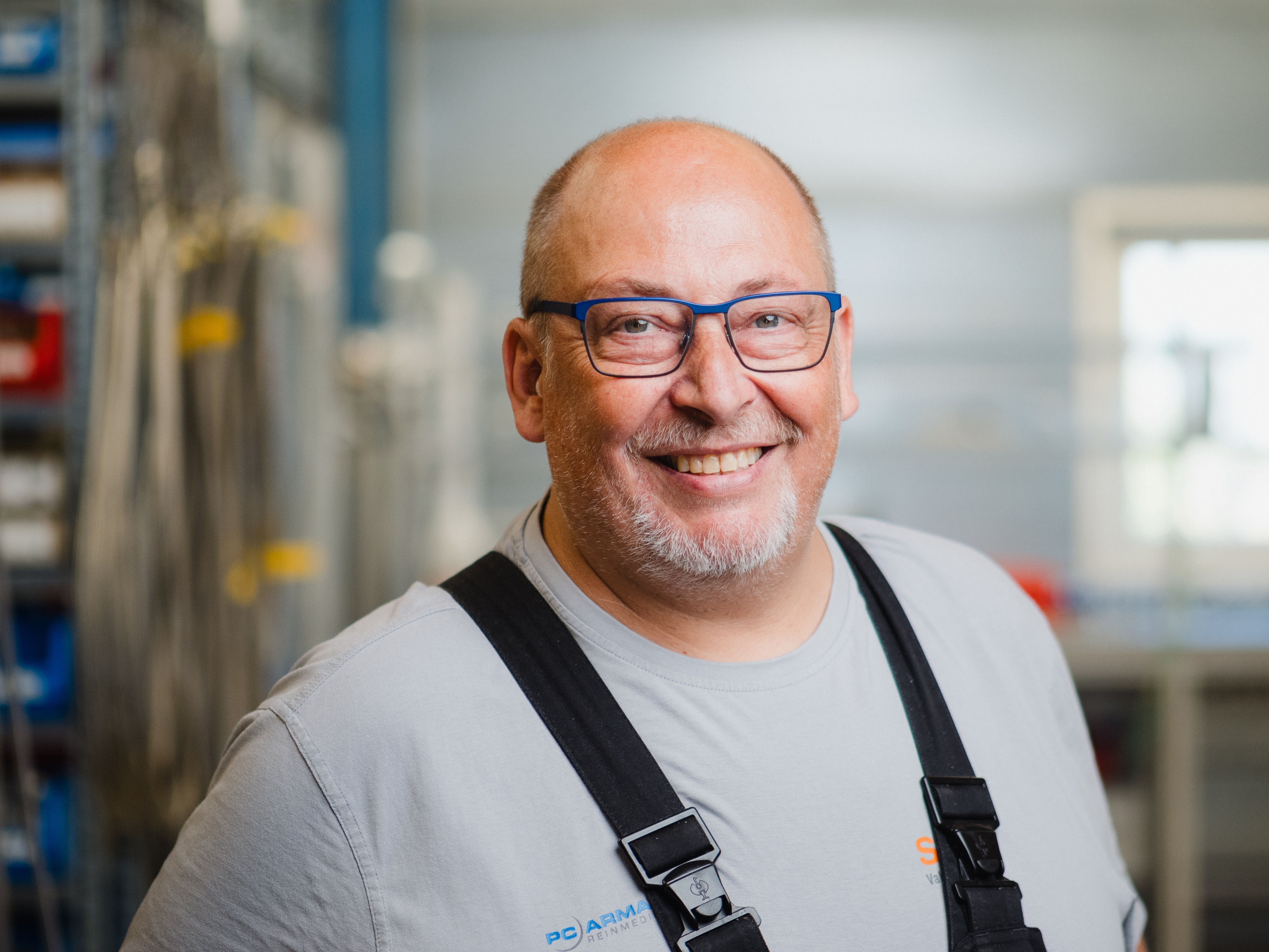 Businessportrait eines Mitarbeiter in der Industrie, der fröhlich lächelt und vetrauensvoll wirkt. Im Hintergrund ist das innere einer Werkhalle zu sehen