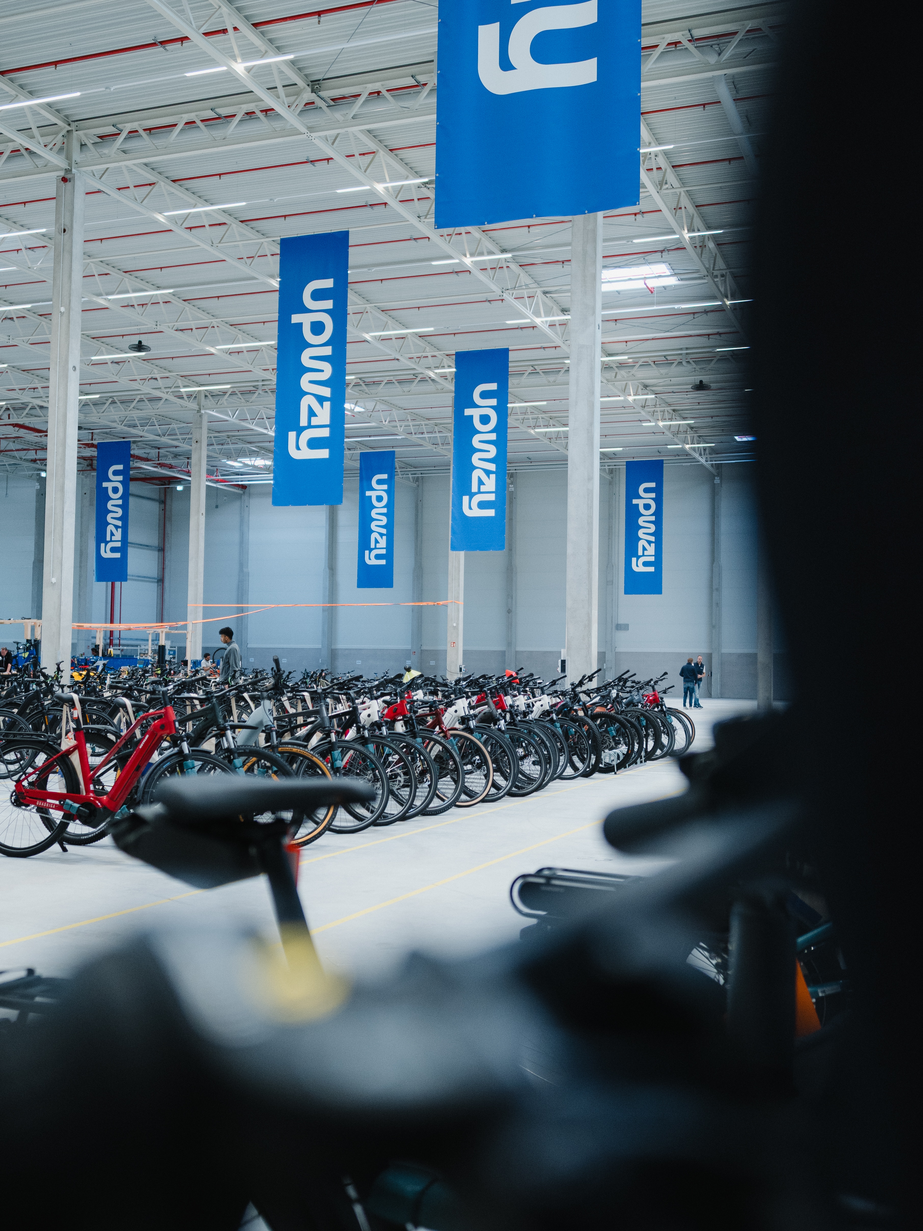 Logistikhalle mit dynamischem Branding: Professionelle Architektur-Fotografie einer E-Bike-Flotte unter blauen Upway-Bannern. Die perspektivische Bildgestaltung unterstreicht die Größe und Innovationskraft des Unternehmensstandorts.