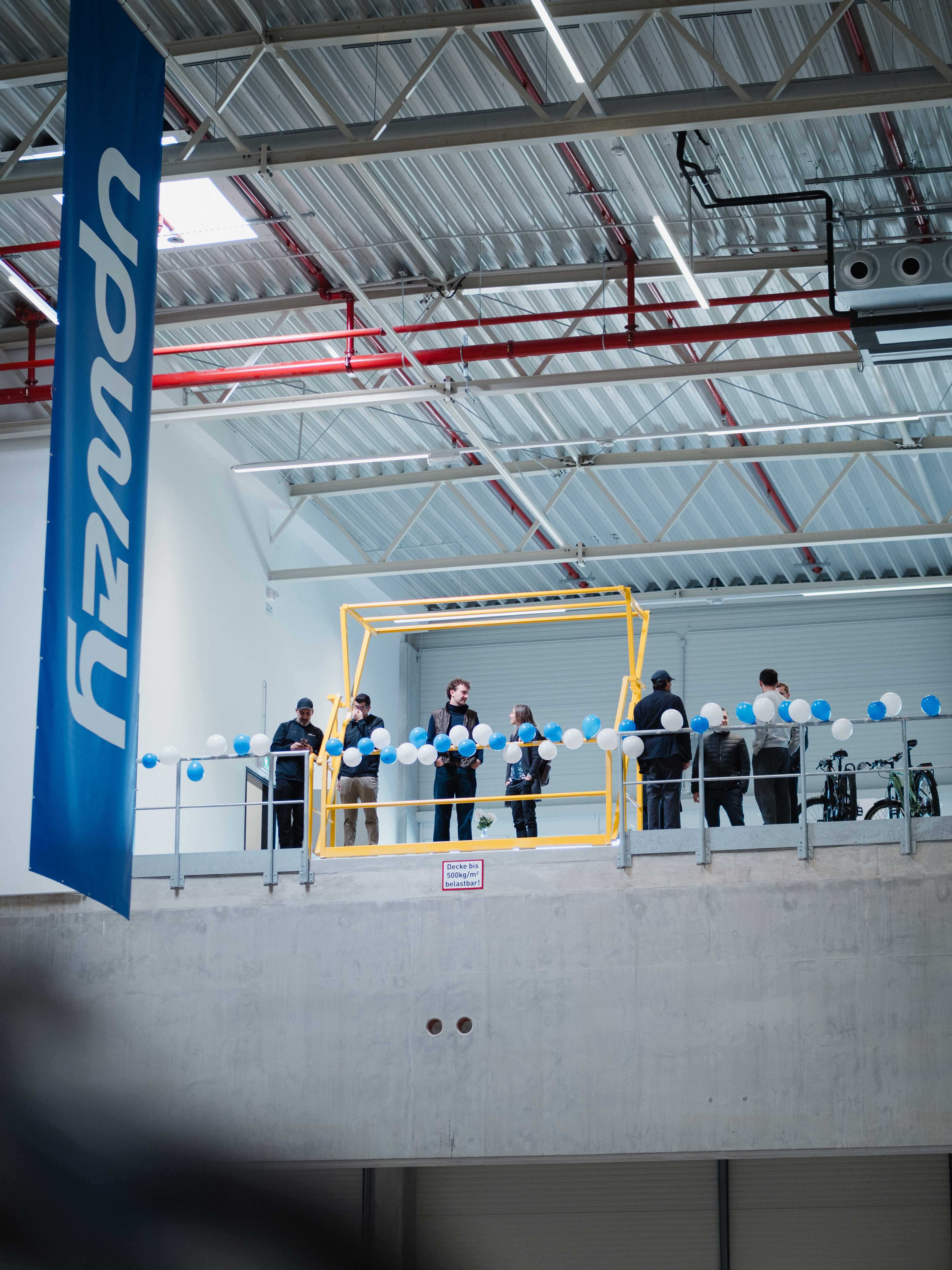Professionelle Event-Fotografie einer Standorteröffnung: Eine Gruppe von Menschen auf der Galerie einer modernen Industriehalle, dekoriert mit feierlichen Ballons und Corporate Branding von Upway. Die weite Perspektive vermittelt Größe und Aufbruchstimmung.