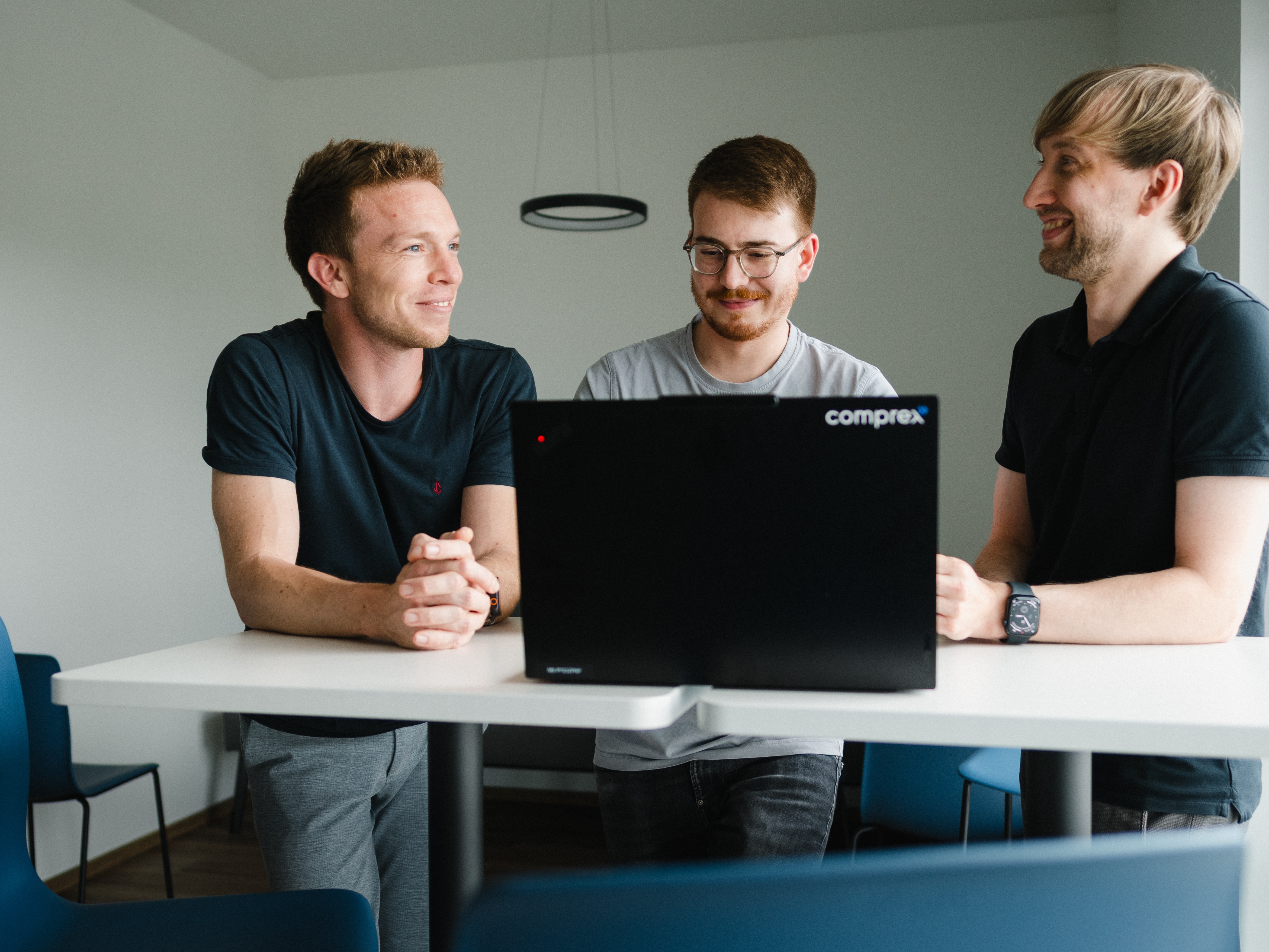Drei Kollegen im lockeren Business-Look tauschen sich gut gelaunt bei der agilen Zusammenarbeit an einem Laptop aus. Modernes Teambild, das flache Hierarchien und zeitgemäße Unternehmenskultur widerspiegelt.